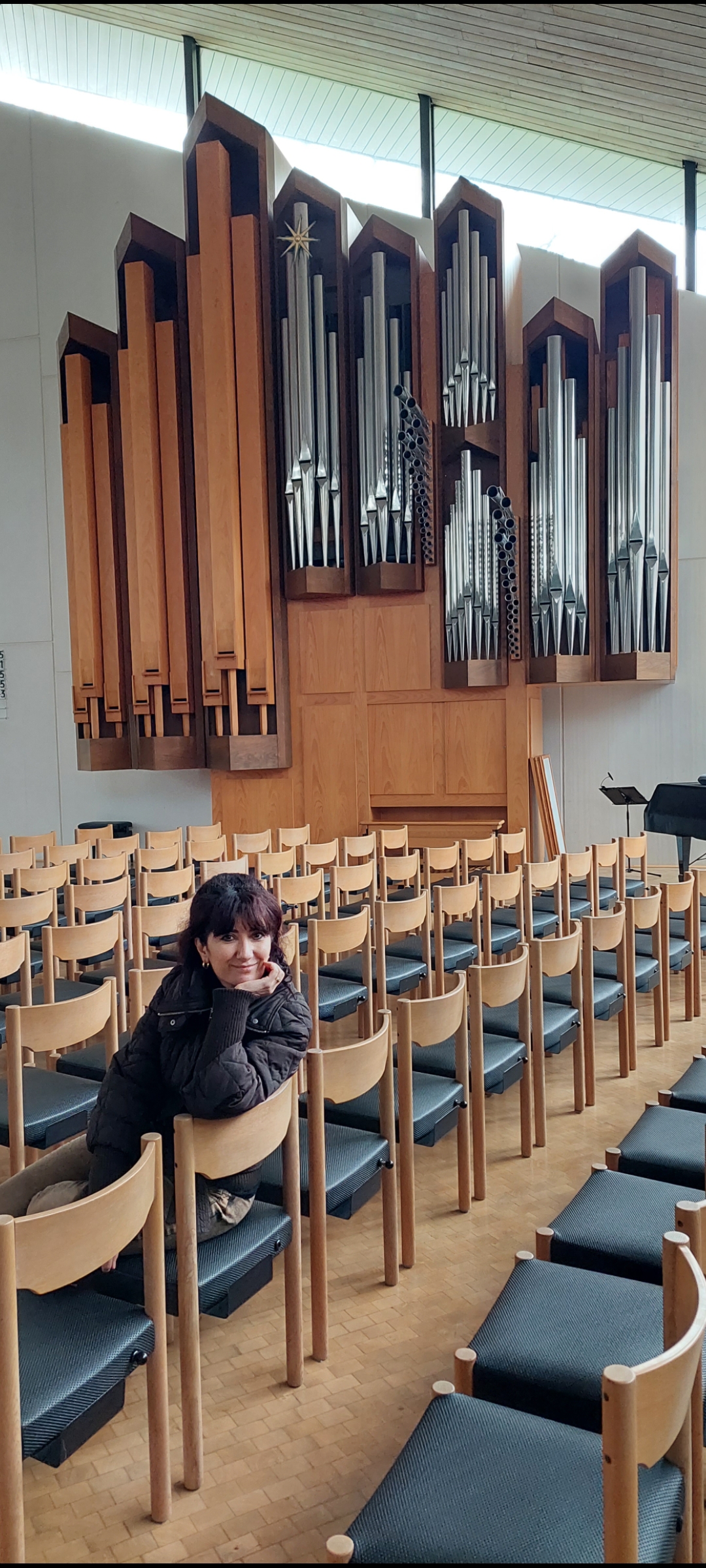 title A woman sits in front of several rows of wooden chairs, near a large pipe organ. A woman sits in front of a large pipe org...