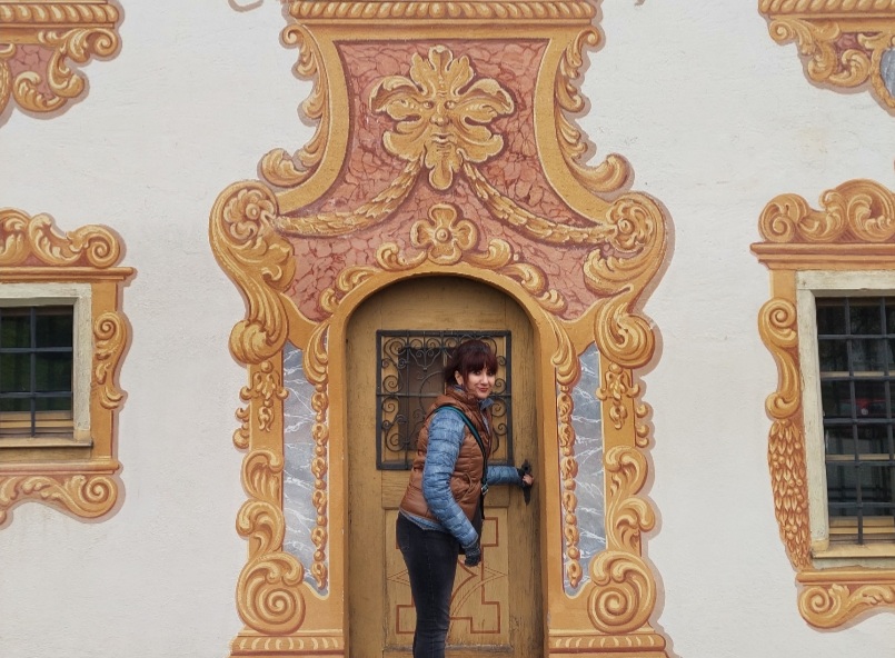 A woman in a blue and brown vest and pants is standing in front of an ornate, gold-framed doorway.
